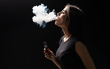 Young beautiful brunette woman smoking, vaping e-cigarette with smoke on black background.